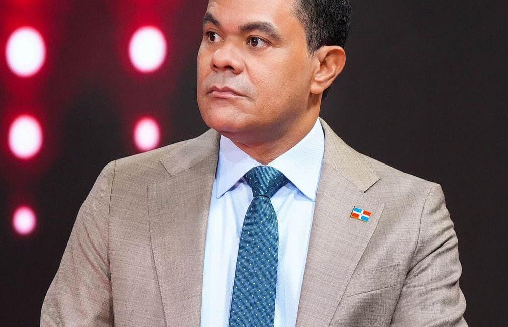 Plano medio del director de la DIDA durante un debate. Usa camisa blanca, corbata azul y un pin de la bandera dominicana. El fondo es un estudio con luces circulares rojas y blancas.