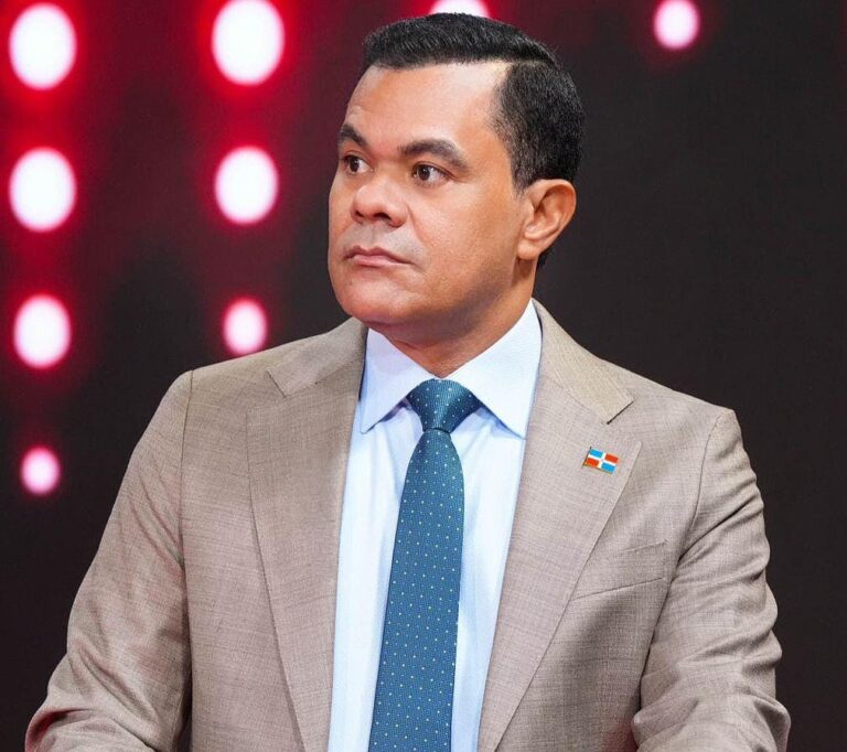 Plano medio del director de la DIDA durante un debate. Usa camisa blanca, corbata azul y un pin de la bandera dominicana. El fondo es un estudio con luces circulares rojas y blancas.