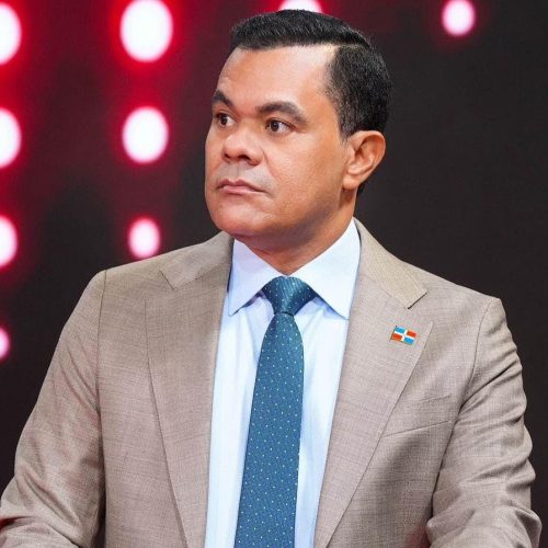 Plano medio del director de la DIDA durante un debate. Usa camisa blanca, corbata azul y un pin de la bandera dominicana. El fondo es un estudio con luces circulares rojas y blancas.